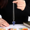 A hand holds a silver crystal necklace with a blue pendant against a black background.