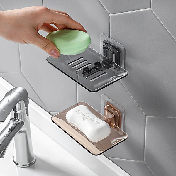 A hand places a green soap onto a clear wall-mounted double layer soap rack next to a faucet.