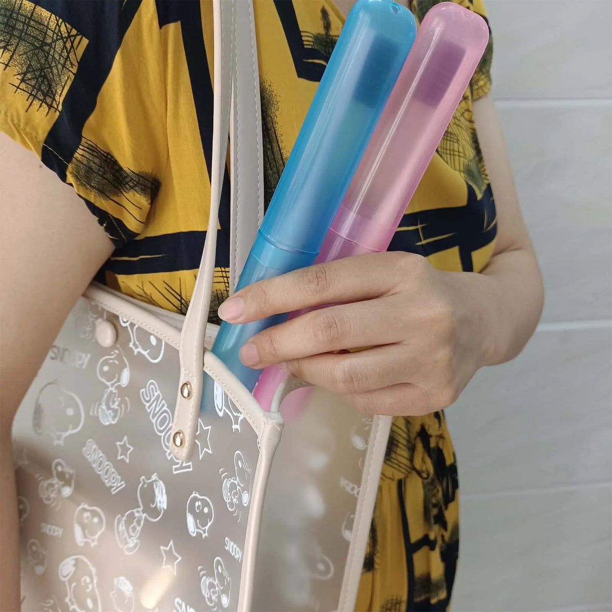 A hand holds a clear plastic toothbrush case with blue and pink bristles against a yellow patterned shirt.