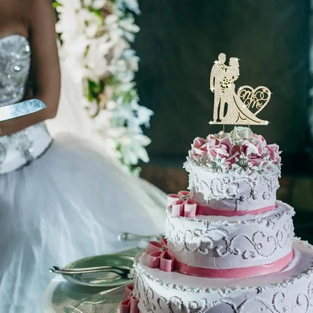 A tiered wedding cake decorated with white frosting and pink flowers, topped with a gold rhinestone Mr & Mrs topper.