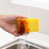 A hand is holding a yellow scouring sponge with a dark, used side, dripping water into a stainless steel sink.