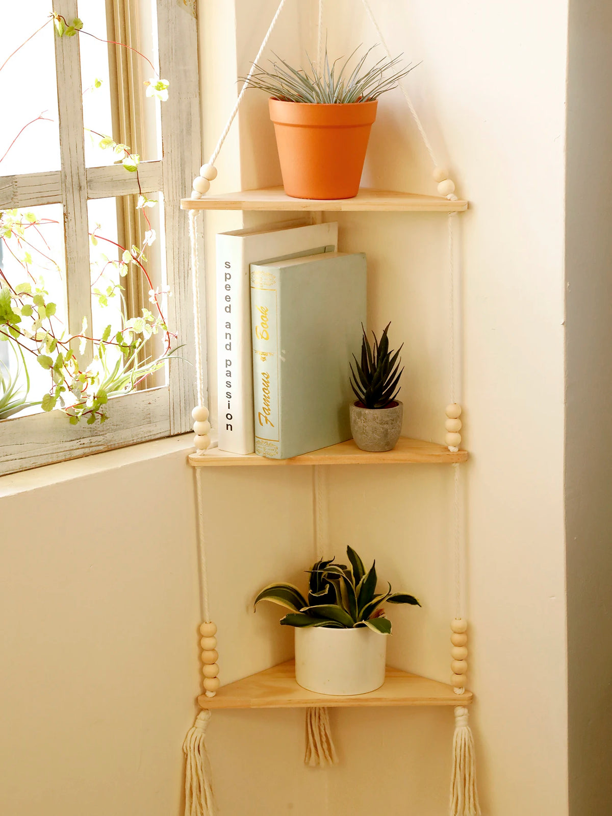 A set of three wooden floating shelves with macrame hangers displaying books and potted plants on a light-colored wall.