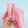 A hand holds a clear glass and a pink microfiber cleaning cloth towel against a light blue background.