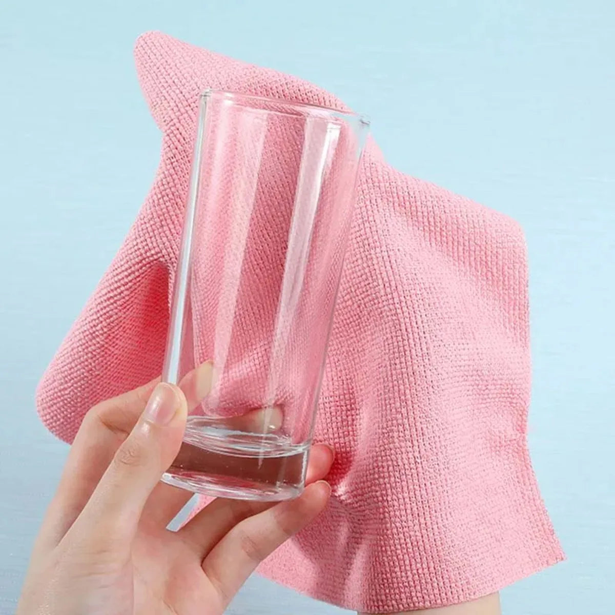A hand holds a clear glass and a pink microfiber cleaning cloth towel against a light blue background.