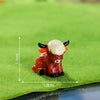 A small, glossy resin figurine of a Highland cow in a reddish-brown color sitting on green grass.