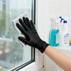 A hand wearing a black nitrile cleaning glove is wiping a window with a white sponge.