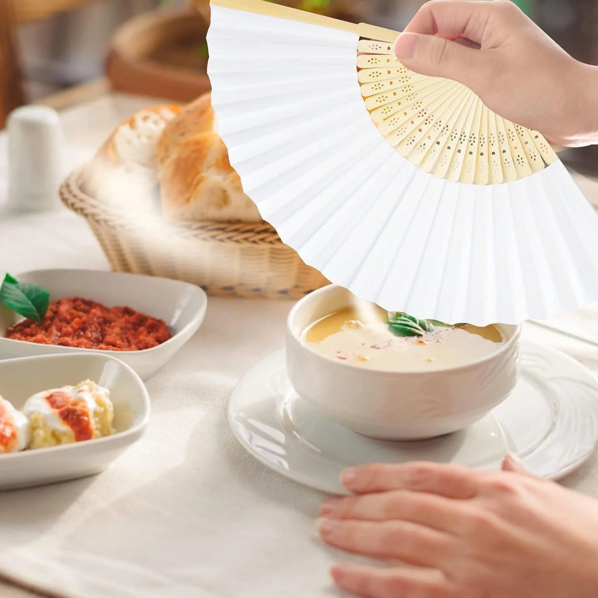 Une main tient un éventail décoratif blanc en bambou au-dessus d'un bol de soupe et de pain.