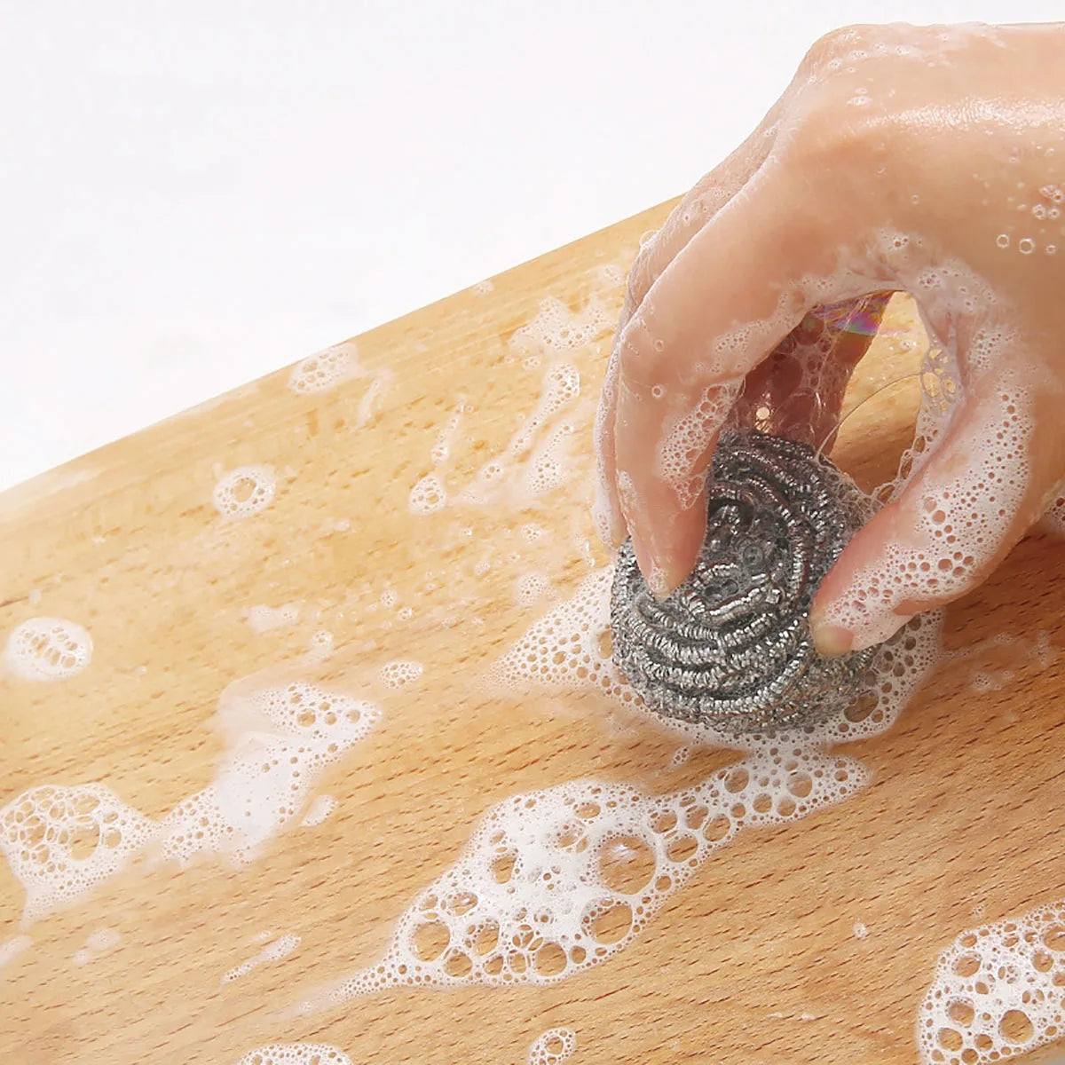 A hand is using a stainless steel scrubber pad to create foam on a wooden surface for cleaning.