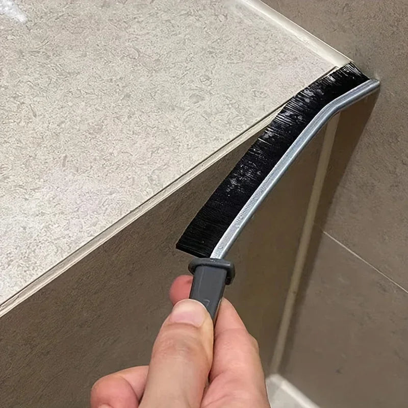 A hand is holding a black hard-bristle crevice cleaning brush, which is attached to a beige cabinet or surface.