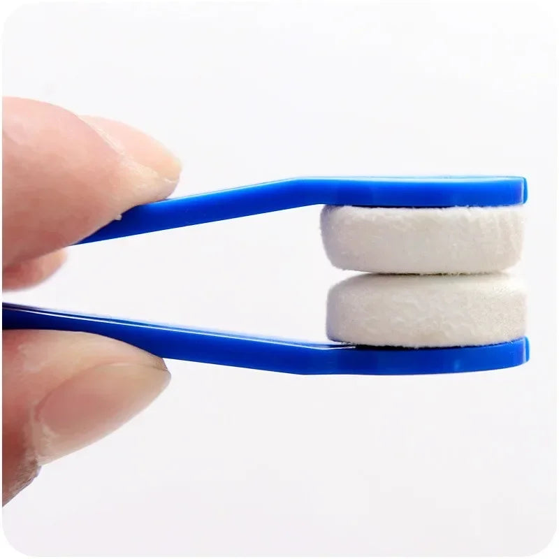 A hand holds a blue cleaning brush with two white microfiber pads for cleaning eyeglasses.