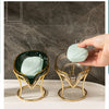 A hand holds a glass soap dish with a light green bar of decorative leaf soap on a gold stand.