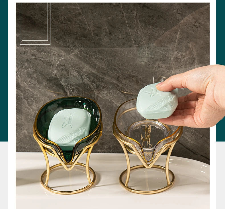 A hand holds a glass soap dish with a light green bar of decorative leaf soap on a gold stand.