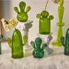 Collection de vases en cactus en verre vert vif, présentés sur des socles blancs et une surface blanche et duveteuse.
