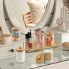 A collection of cosmetic brushes and makeup products stored in glass apothecary jars with wooden lids on a white vanity surfa