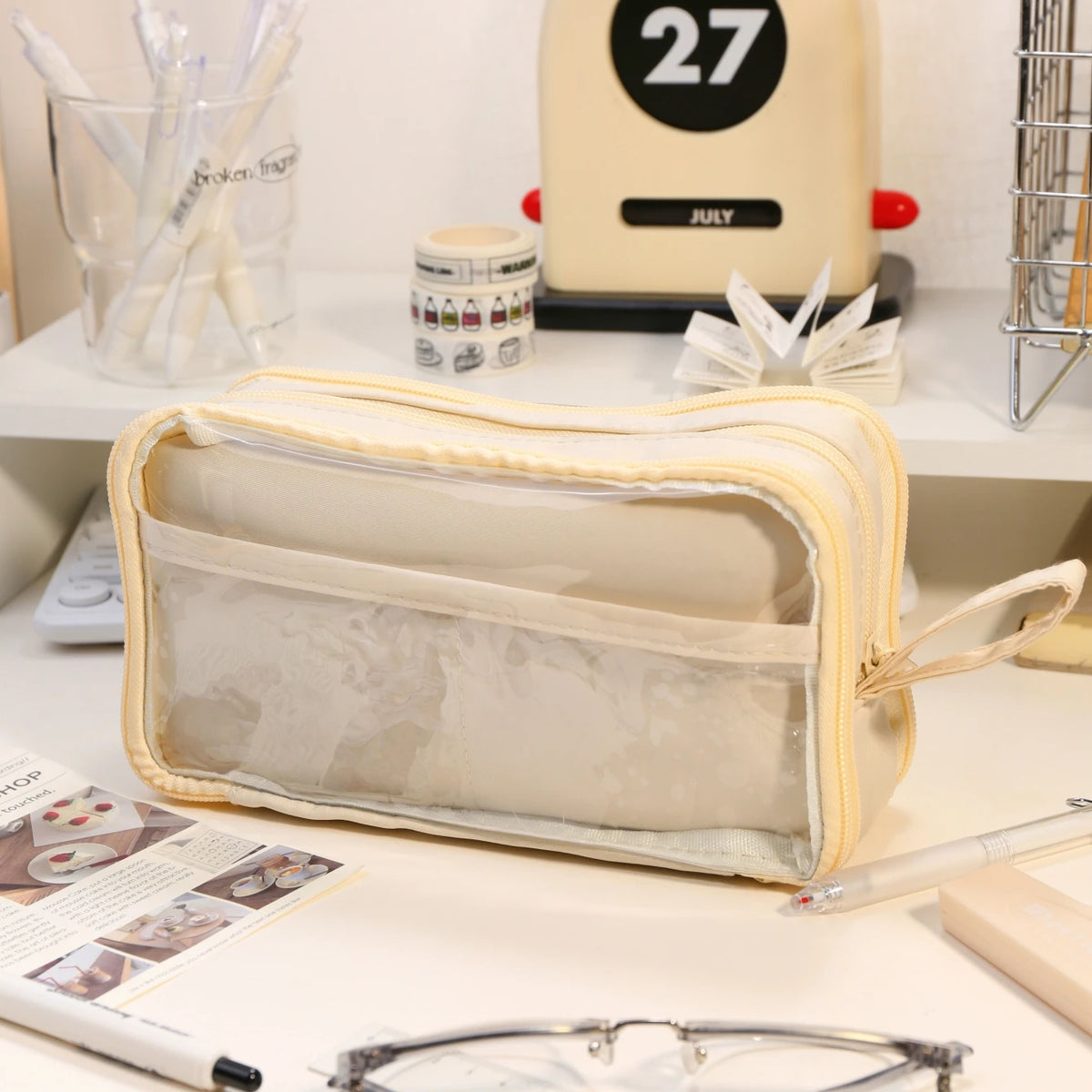 A beige transparent pencil pouch is displayed on a white surface next to a yellow appliance and other stationery.