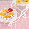 A white plastic dessert stand holds small cups filled with fruit and candy on a pink checkered surface.