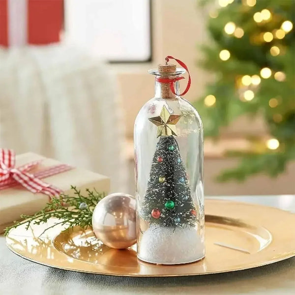 A clear glass message bottle decorated with a Christmas tree and sparkling glitter sits on a gold plate near a festive green 
