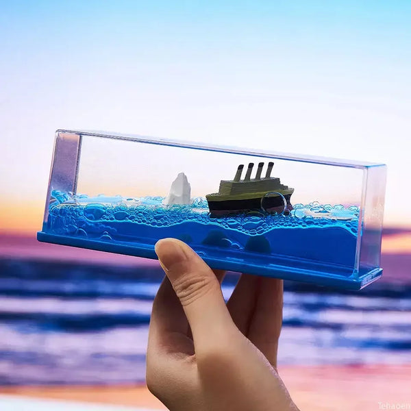 A hand holds a clear glass box containing a miniature ship ornament submerged in blue water.