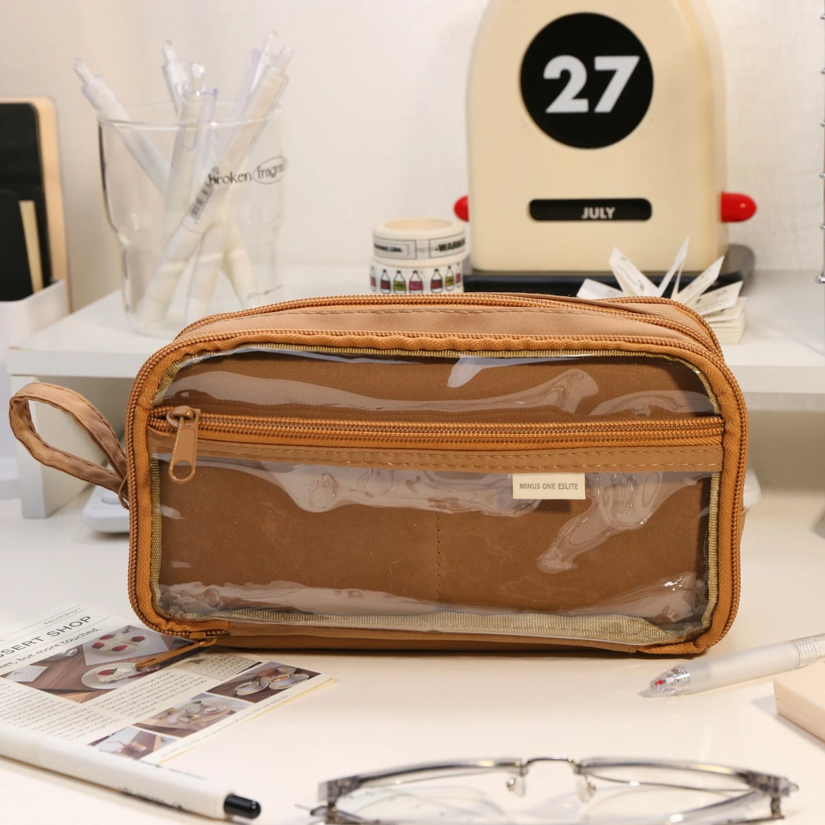 A brown transparent pencil pouch is shown on a white surface next to various writing tools and accessories.