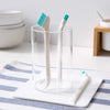 A clear glass filled with a white cleaning brush set featuring two white and one teal silicone brushes resting on a white tra