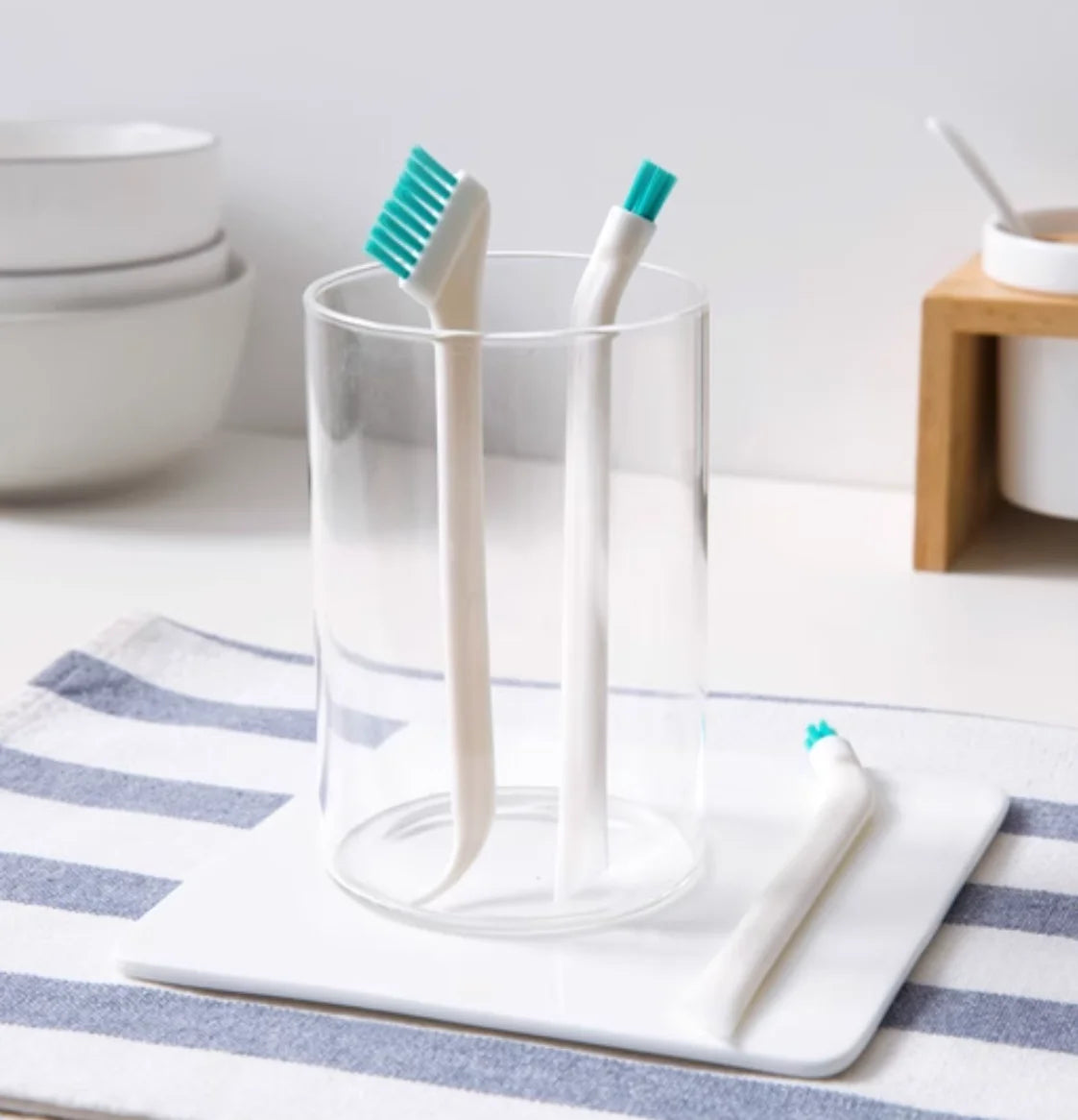 A clear glass filled with a white cleaning brush set featuring two white and one teal silicone brushes resting on a white tra