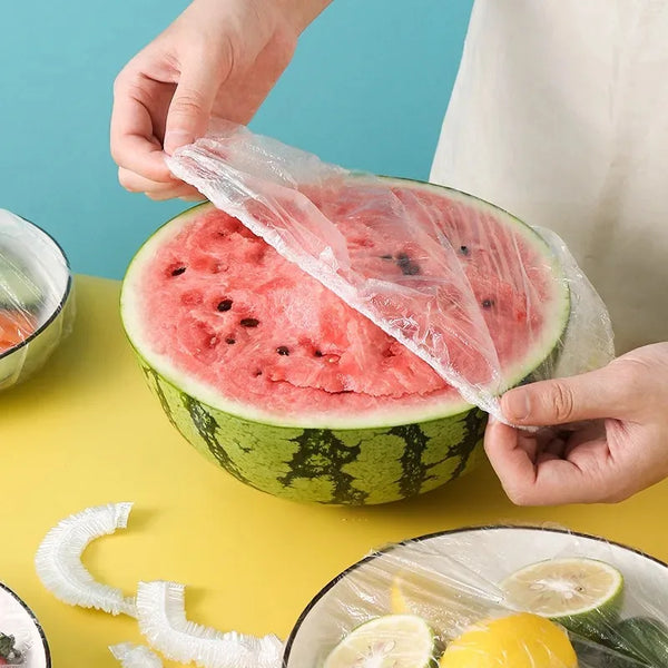 Hands are placing a clear plastic food cover over a slice of watermelon on a yellow surface.