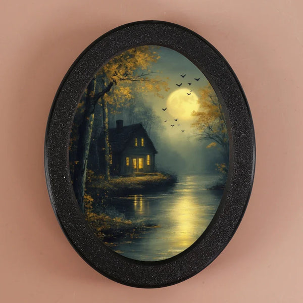 An oval photo frame featuring a moody landscape of trees, water, and a bright full moon with small birds flying.