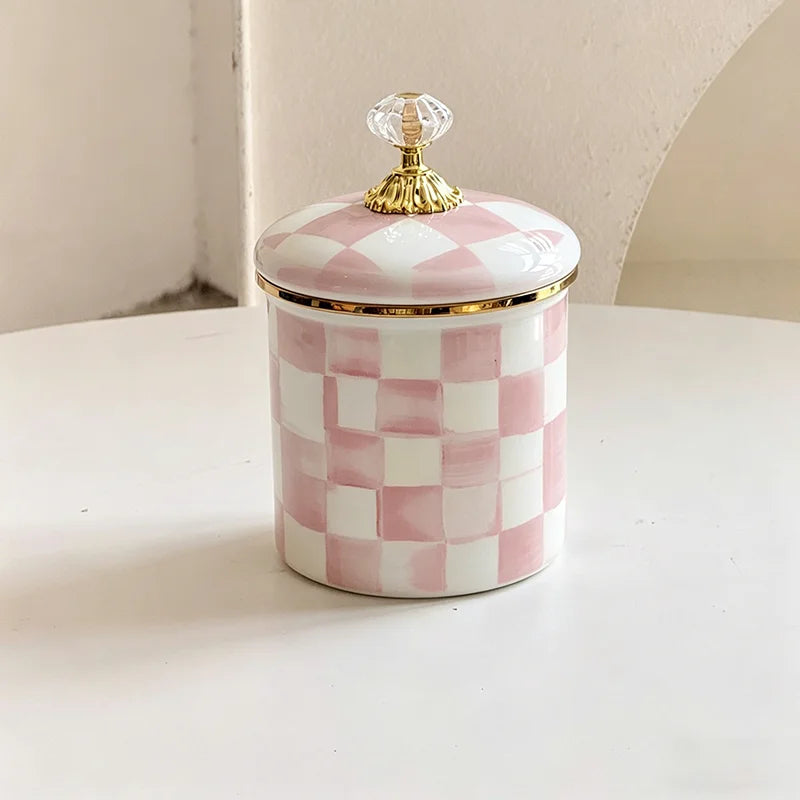 A white and pink checkered enamel storage jar with a gold-toned lid sits on a white surface.