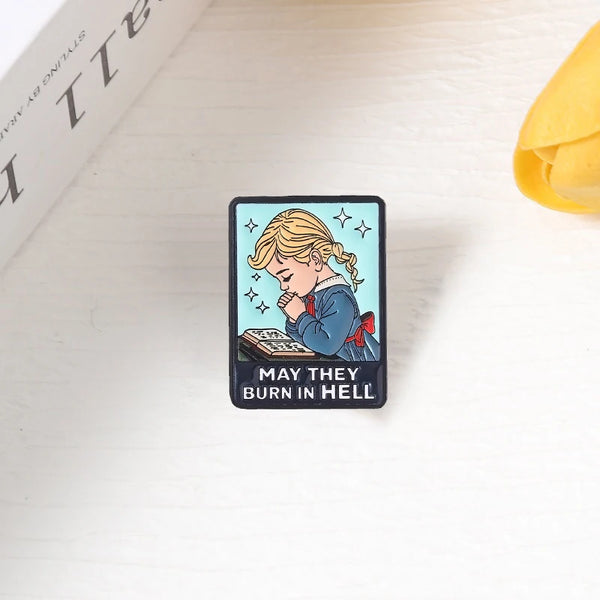 A black enamel lapel pin featuring a cartoon illustration of a girl looking distressed with the text May They Burn in Hell.