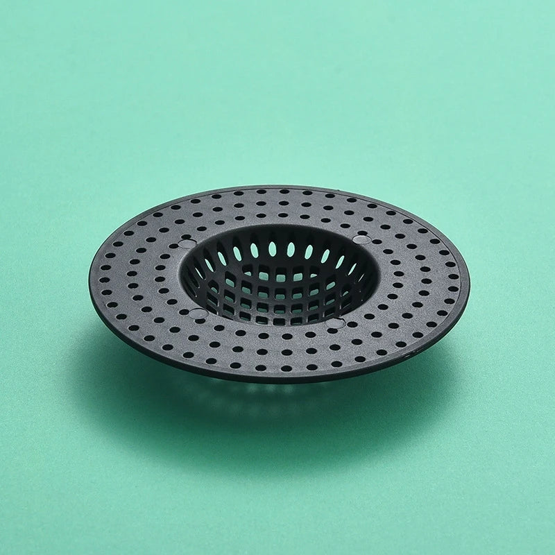 A black kitchen sink drain strainer with a perforated mesh design, photographed on a light green surface.