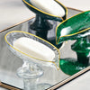 A clear glass oval soap dish with drainage holes holds a white bar of soap and is displayed in a square tray.