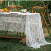 A white lace retro tablecloth is set on an outdoor table surrounded by greenery and wooden chairs.