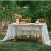 A rustic outdoor table set with a lace tablecloth, wooden chairs, and floral decorations in a garden setting.