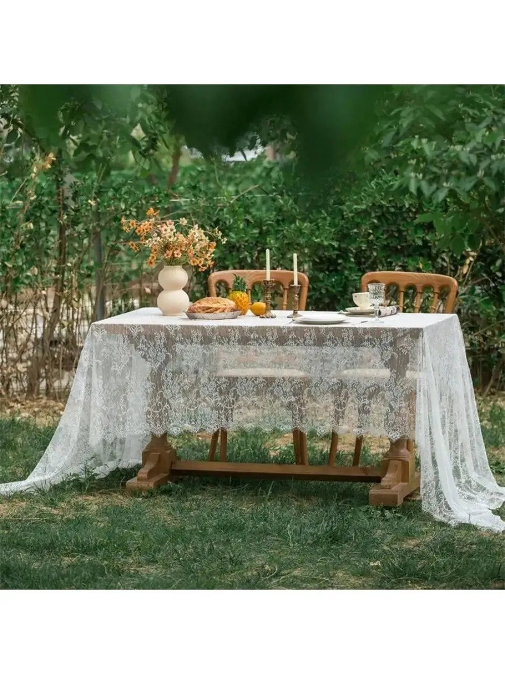 A rustic outdoor table set with a lace tablecloth, wooden chairs, and floral decorations in a garden setting.