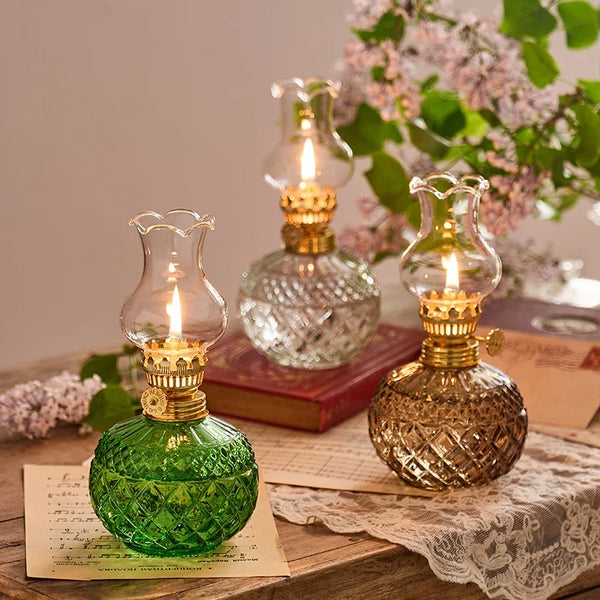 A vintage rustic oil lamp in green glass sits on a wooden surface next to a stack of paper and pink flowers.