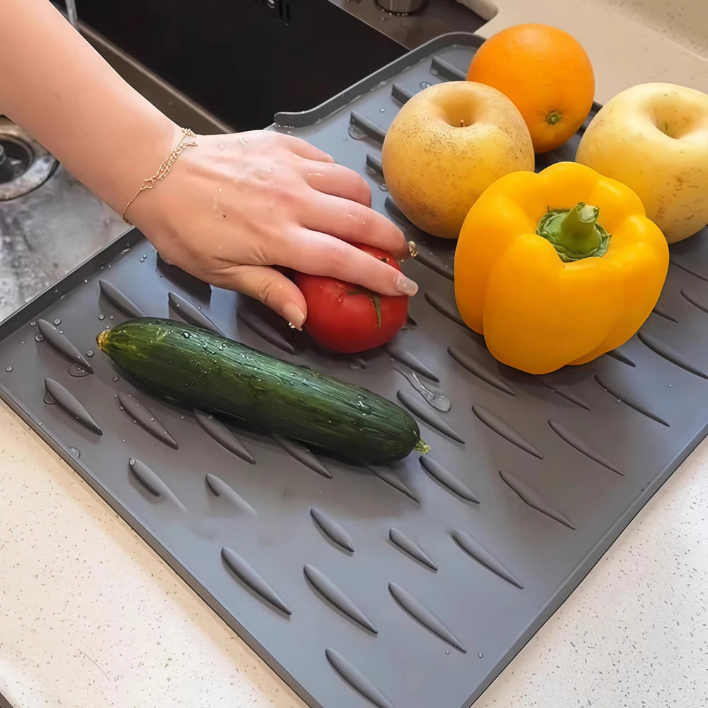 A hand places various fresh produce like apples, a bell pepper, and a cucumber onto a silicone drain mat.