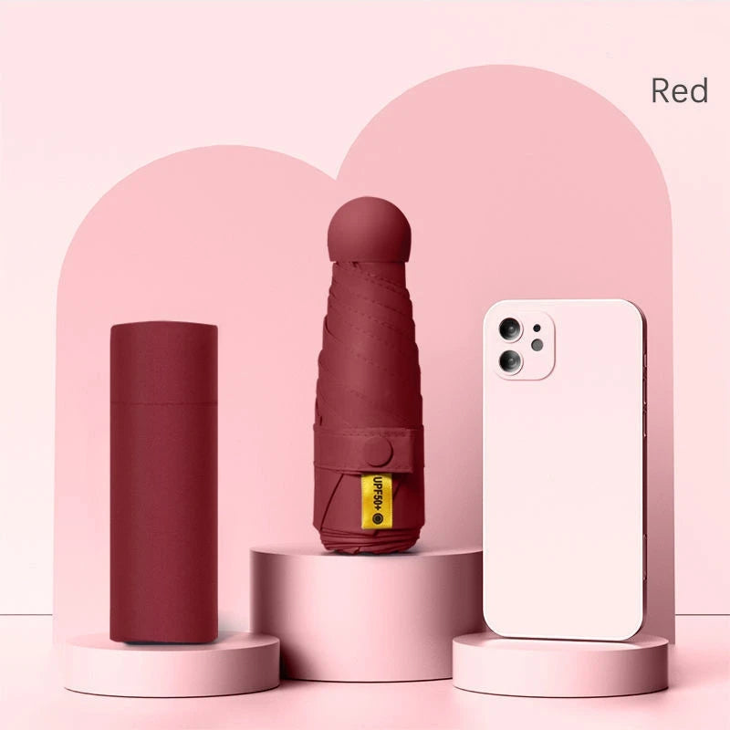 A maroon folding UV protection umbrella standing next to a white smartphone in a pink display box.