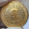 Runder goldener Münze mit stilisiertem Schädeldesign, umgeben von verzierten Schnörkeln.