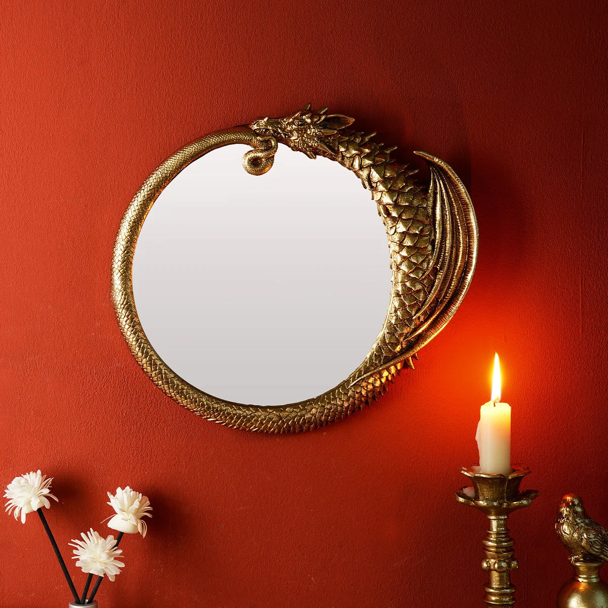 A round, ornate gold-framed mirror hangs on a terracotta red wall next to a lit candle and white flowers.