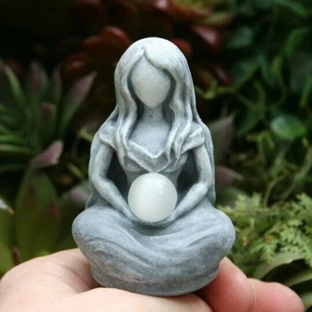 A hand holds a gray resin statue of a female figure with long hair and a round white orb, set against a blurred green backgro
