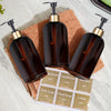 A collection of dark amber refillable pump soap dispenser bottles and a paper label are displayed on a light surface.