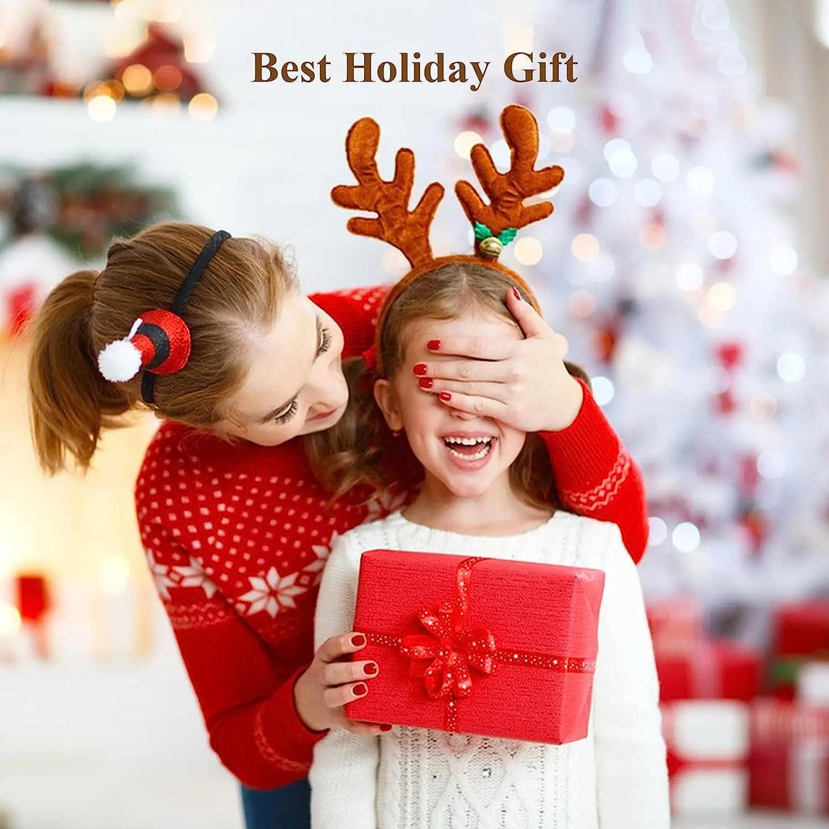 A young girl is playfully covering her eyes with reindeer antlers while holding a red gift box in front of a blurred Christma