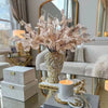 A large arrangement of dried pink and white flowers in a gold vase displayed on a glass table next to a sofa.
