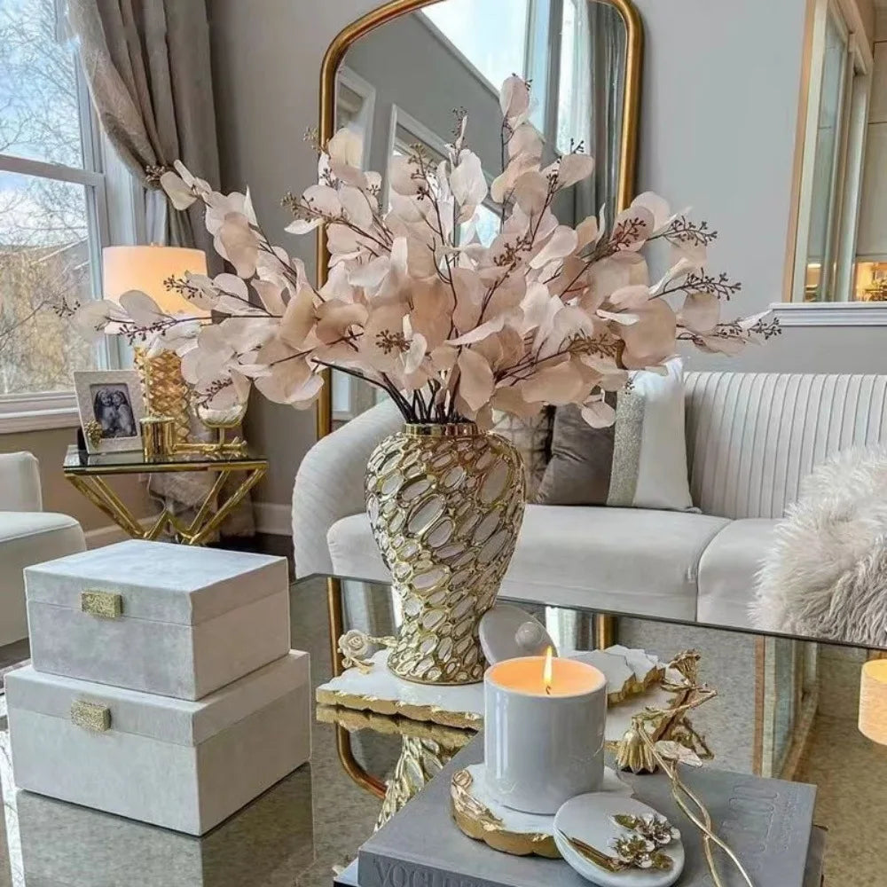 A large arrangement of dried pink and white flowers in a gold vase displayed on a glass table next to a sofa.