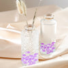 Glass jars filled with purple crushed ice stones and dried floral elements on a beige fabric.