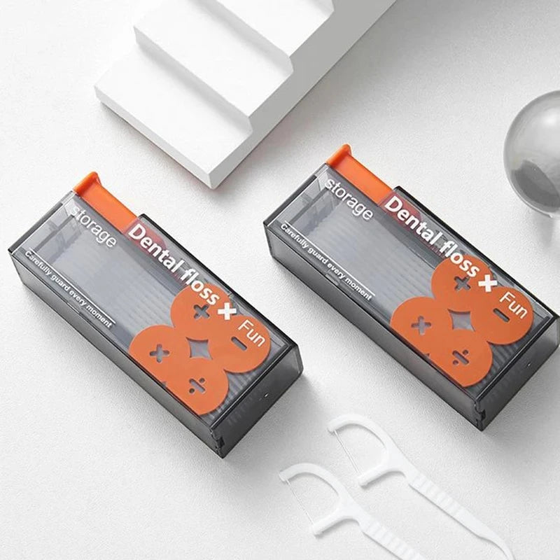 Dispenser portátil de hilo dental negro con detalles naranjas y palitos reutilizables sobre una superficie blanca.