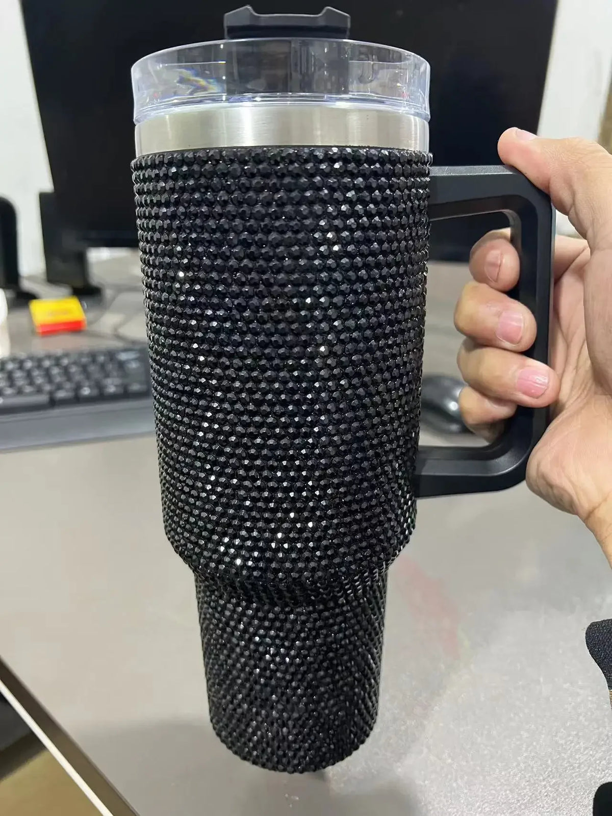 Una mano sostiene un termo negro aislado con acabado de strass texturizado y tapa transparente.