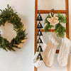A vintage metal wind chime decoration hangs on a wooden rack, surrounded by green foliage and white fabric.