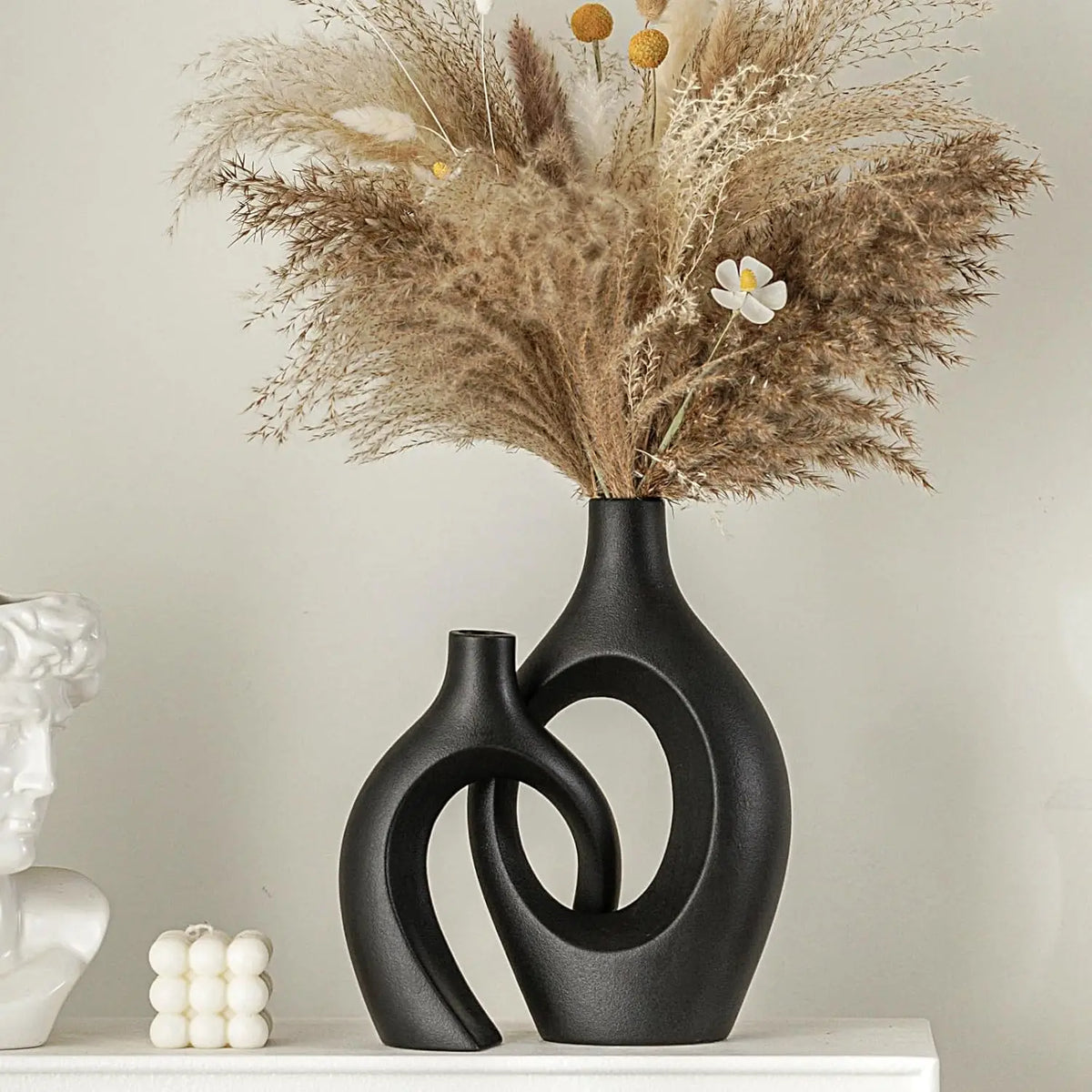 A black ceramic vase holds dried grasses and a white flower on a white shelf.