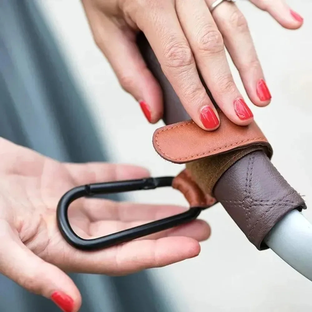 A hand is holding a brown leather stroller hook carabiner accessory.